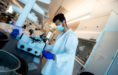 researcher wearing white lab coat and face mask in a lab overlooking lab equipment